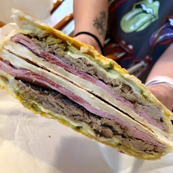 A person holding a large, sliced Cubano sandwich, revealing layers of ham, pork, cheese, and pickles.