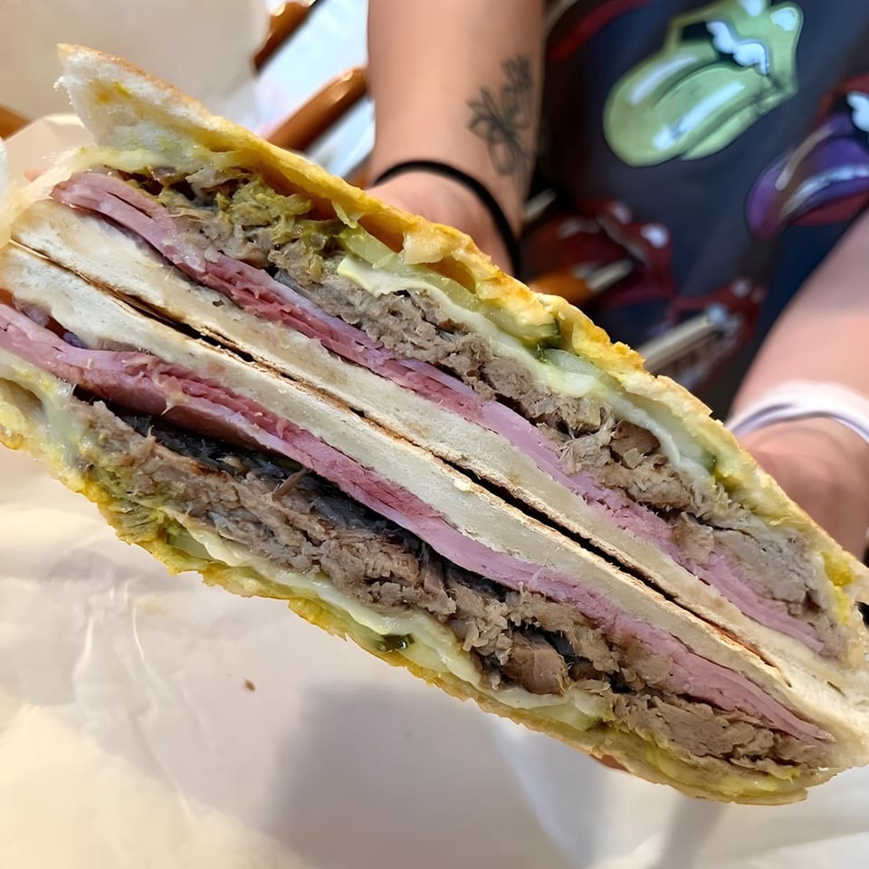 A person holding a large, sliced Cubano sandwich, revealing layers of ham, pork, cheese, and pickles.