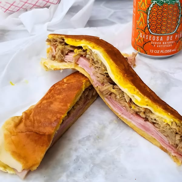 A Cubano Bros Cuban sandwich, cut in half, with a pineapple soda on white paper.