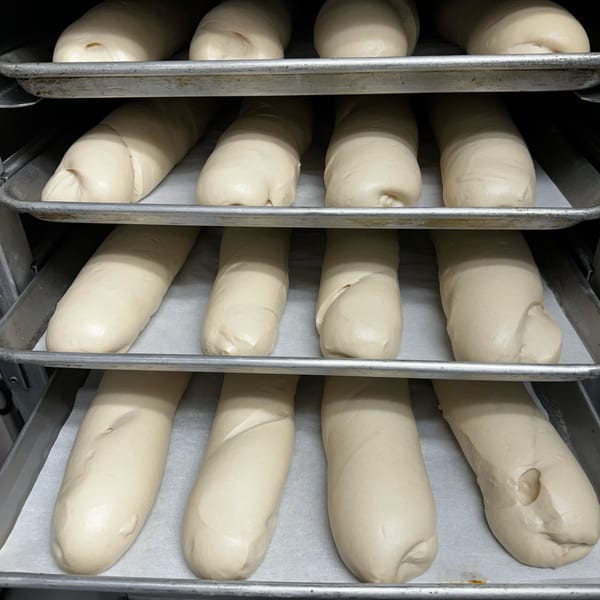 Rows of unbaked Cuban bread dough loaves resting on baking sheets in a metal rack.