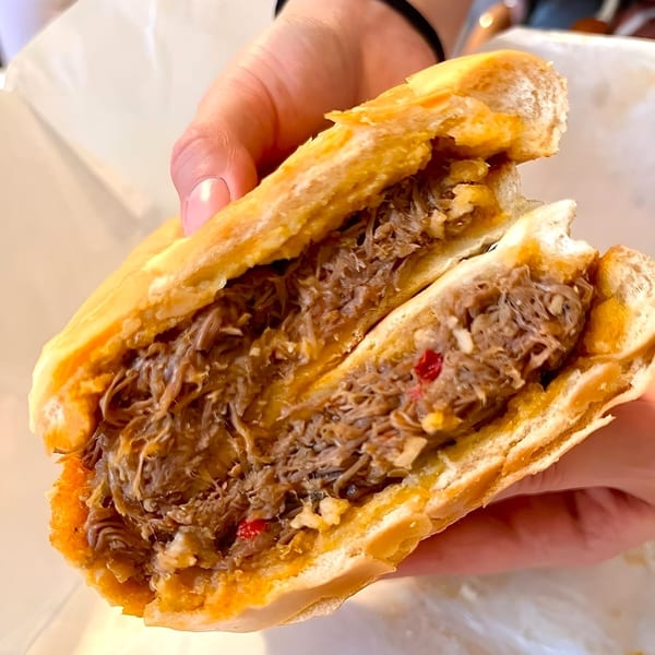 A hand holds a large, open Cubano Bros sandwich, revealing generous shredded meat filling.