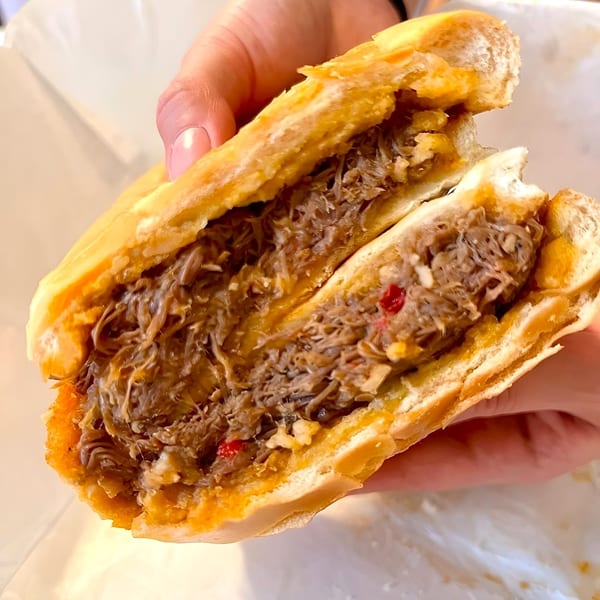 A hand holds a large, open Cuban sandwich overflowing with shredded meat and red peppers.