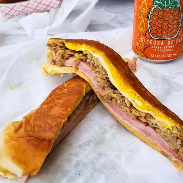 A Cubano Bros Cuban sandwich, cut in half, with a pineapple soda can on white paper.